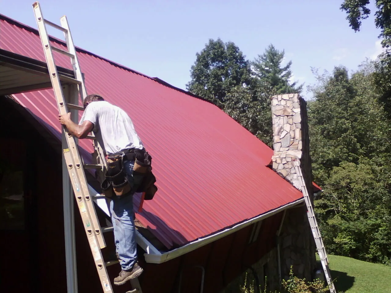 Roofer on a ladder beside a red metal roof and stone chimney — Soffit & Fascia Repair by Roofing Brothers in Zanesville